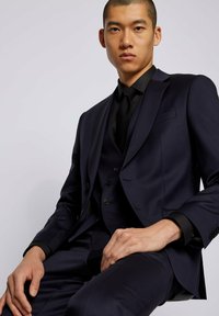 Granatowy, dopasowany garnitur z wąskim krojem, warstwową kamizelką i czarną koszulą. Garnitur ma eleganckie klapy i guziki zdobnicze.