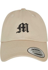 Bež puuvillane müts kumerate äärega ja reguleeritava rihmaga. Esiküljel on musta värvi broderitud "M" logo. Tekstureeritud kangas.