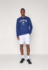 Marineblå Adidas langærmet sweatshirt med hvidt logo, parret med hvide shorts. Sorte træningssko og ankelstrømper fuldender outfittet.