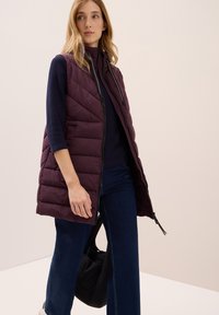 Burgundský vatovaný vest s uzavřením na zip a designem ve tvaru šípů, nošený přes tmavý svetr a spárovaný s tmavými zvonovými džíny. Jednoduchá černá taška.