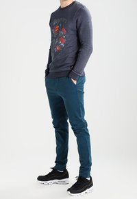 Felpa blu scuro con grafica floreale e testo; abbinata a pantaloni turchesi e scarpe sportive nere con suola bianca.