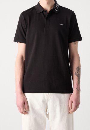 Mand iført en sort kortærmet poloshirt med hvid tekst på kraven og lyse bukser, der viser tatoveringer på sin højre underarm.