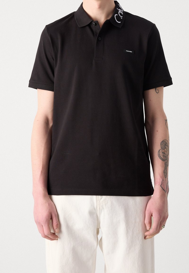 Homme portant un polo noir à manches courtes avec un texte blanc sur le col et un pantalon de couleur claire, montrant des tatouages sur son avant-bras droit.