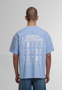 Tricou din bumbac de culoare albastru deschis, cu text grafic alb și ilustrații ale unor tetrapackuri de film pe spate. Mâneci scurte și croială lejeră.