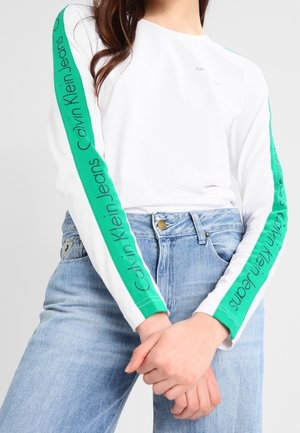 Weißes Langarmoberteil mit grünen Streifen an den Ärmeln mit dem Schriftzug „Calvin Klein Jeans“, kombiniert mit hellblauen Jeans, Hände verschränkt.