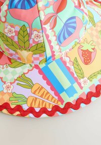 Chapeau de soleil coloré avec un mélange de motifs floraux et végétaux en rose, vert et bleu. Bord rouge ondulé et tissu doux.