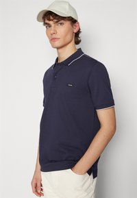 Polo navy de algodón con cuello y acentos de manga en blanco. Presenta un pequeño parche de logo en el pecho. Combinado con pantalones de color claro.