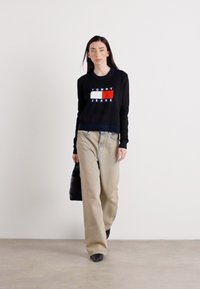 Une femme marche avec confiance dans une chemise noire à manches longues avec un logo, associée à un pantalon clair et un sac à main noir, sur un fond neutre.