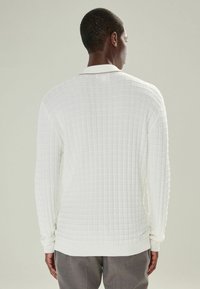 Un homme de dos porte un pull long à col texturé blanc et un pantalon gris sur un fond clair uni.