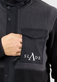 Giacca in pile nera con chiusura con zip, bottoni a pressione e una tasca sul petto con un logo bianco. La texture è morbida e lanosa.