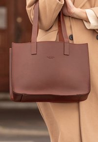 Personne dans un manteau beige tenant un sac fourre-tout en cuir marron avec deux anses et un logo embossé subtil à l'avant.