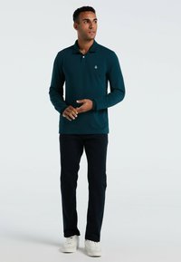 Lange mouwen teal polo shirt met een kraag en knoopsluiting, gecombineerd met donkere broek en witte sneakers. Subtiel logo op de borst.
