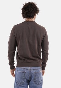 Homme aux cheveux bouclés portant une chemise marron à manches longues et un jean bleu, debout de dos sur un fond uni.