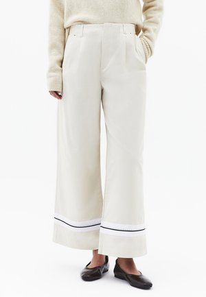 Kremowe spodnie o szerokich nogawkach z biało-czarnym obszyciem przy krawędzi, zestawione z beżowym swetrem i czarnymi płaskimi butami.
