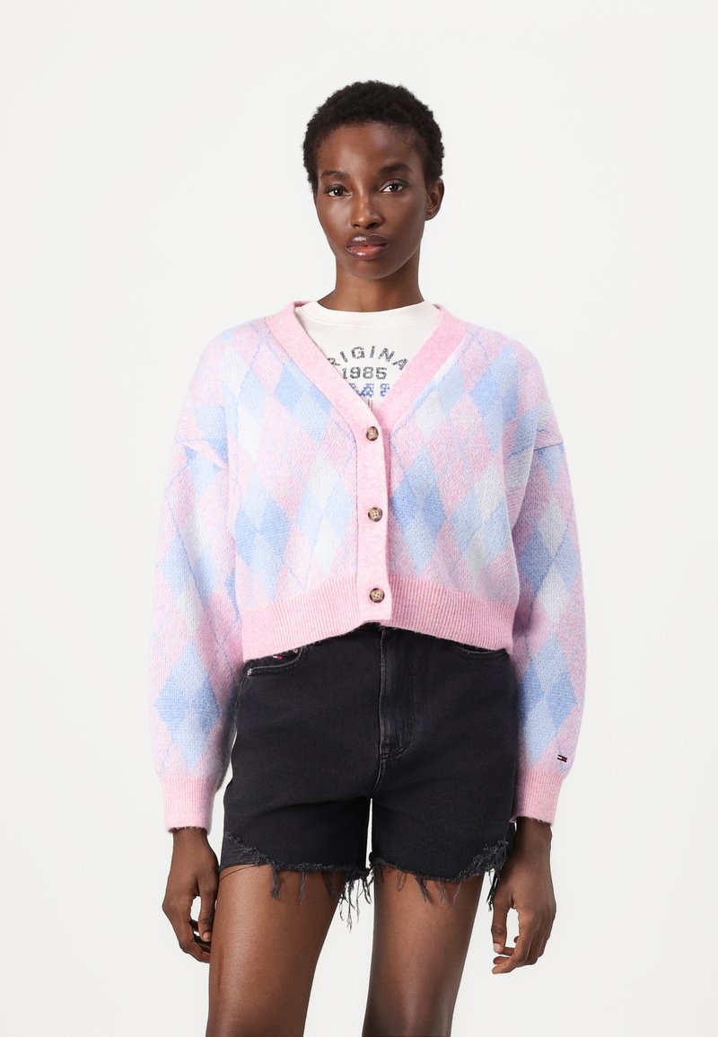 Cárdigan de patrón argyle en rosa y azul, estilo corto, con cierre de botones, textura suave, combinado con pantalones cortos desgastados en negro.