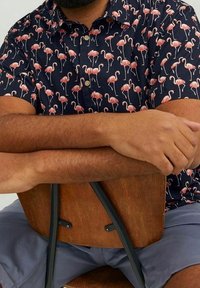 Camisa de manga corta en azul marino con un patrón de flamencos, que presenta botones y un cuello de punta. Los brazos del modelo descansan sobre una silla de madera.