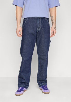 Pantaloni cargo din denim albastru închis, cu picioare drepte, având buzunare laterale și cusături contrastante. Asortați cu pantofi violeți.