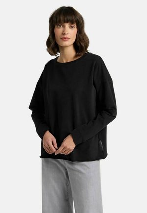 Frau mit kurzen dunklen Haaren, die ein lockeres schwarzes Langarmshirt und hellgraue, weit geschnittene Hosen trägt, steht vor einem einfarbigen Hintergrund.