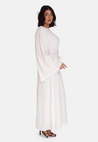 Robe blanche longue avec de larges manches évasées, taille cintrée et jupe fluide. Tissu lisse avec un éclat subtil, associé à des talons aiguilles noirs à lanières.