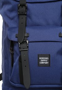 Sac à dos en tissu bleu avec des sangles à boucle noires et une poche avant étiquetée « Herschel Supply Company ».