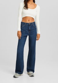 Jeans svasati blu scuro con vita alta, cuciture dettagliate e tasche frontali, abbinati a un top aderente a maniche lunghe bianco a coste.