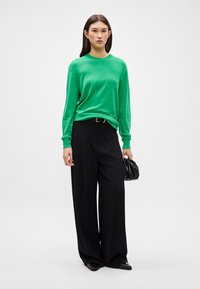 Πράσινο μακρυμάνικο πουλόβερ συνδυασμένο με μαύρες φαρδιές παντελόνες. Το σύνολο διαθέτει άνετη εφαρμογή και υφή πλεκτού με μια κομψή μαύρη τσάντα.