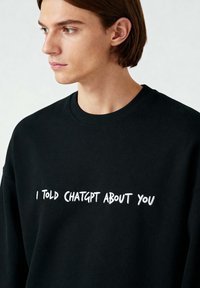 Felpa nera con scollatura tonda. Presenta un testo ricamato bianco: "HO PARLATO DI TE CON CHATGPT." Texture morbida e vestibilità rilassata.