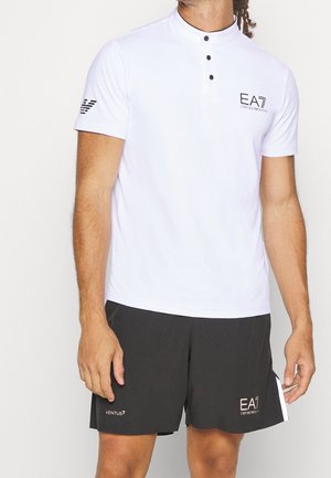 Männlich, trägt ein weißes, kurzärmliges Hemd mit Knopfleiste und schwarzen Shorts mit EA7 Emporio Armani-Logos, steht vor einem einfarbigen Hintergrund.