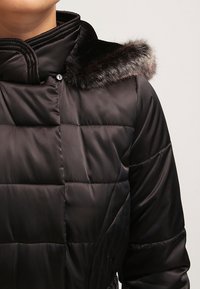 Černá péřová bunda s prošívaným designem a límcem z umělé kožešiny. Má vysoký límec a hladký, lesklý povrch. Tlačítka na přední straně.