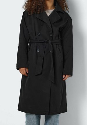 Personne portant un trench-coat noir à double boutonnage avec ceinture nouée, manches longues et poches, associé à un jean bleu clair.