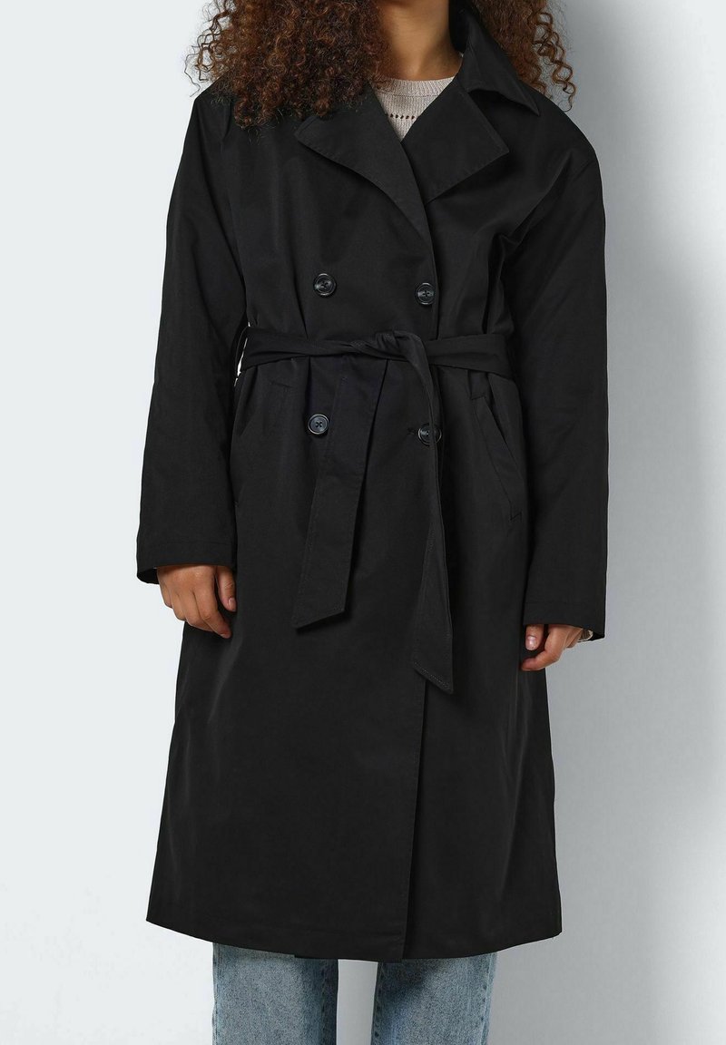Personne portant un trench-coat noir à double boutonnage avec ceinture nouée, manches longues et poches, associé à un jean bleu clair.