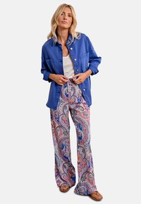 Camicia blu a maniche lunghe, canotta bianca, pantaloni larghi a fantasia paisley colorati e sandali marroni. I tessuti sono leggeri e rilassati.