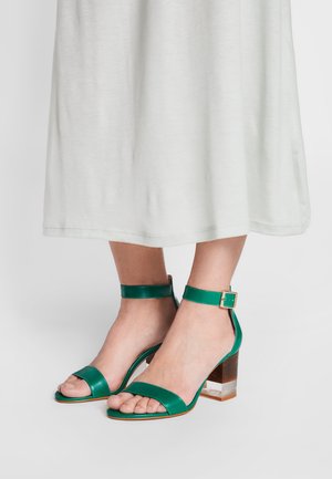 Sandalias de tacón verdes de cuero con correas en el tobillo, puntera abierta y tacón de bloque transparente. Textura suave y diseño minimalista.