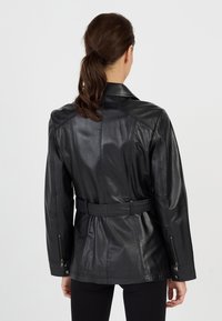 Kobieta z brązowymi włosami związanymi w kucyk, ubrana w czarną skórzaną kurtkę z paskiem i czarne spodnie, pokazana z tyłu na jednolitym tle.