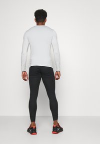 Pitkähihainen, vaaleanharmaa urheiluyläosa, mustat tiukat leggingsit ja mustat lenkkarit punaisilla yksityiskohdilla, nähtynä takaa.