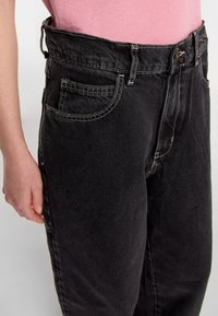 Black denim jeans with a high waist, large side pockets, and white stitching details. The fabric has a smooth texture.