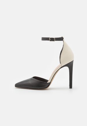 Chaussure à talon haut noire et beige avec un embout pointu, une sangle à la cheville, et des matériaux contrastés. Présentant un design élégant et une texture lisse.