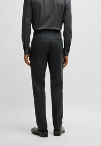 Pantaloni neri slim-fit con una texture liscia, dotati di due tasche posteriori con bottoni e dettagli di pieghe sottili, abbinati a scarpe nere.