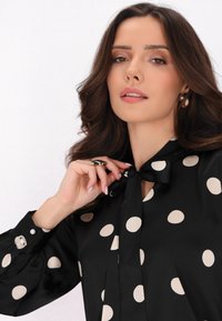 Blusa nera con grandi pois beige, collo a fiocco e maniche lunghe. Presenta polsini con bottoni e una texture di tessuto liscia.