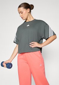 Γκρίζο κοντομάνικο μπλουζάκι με λευκές τρεις ρίγες στα μανίκια, συνδυασμένο με χαλαρές ροζ παντελόνες προπόνησης που διαθέτουν μικρό λευκό λογότυπο. Κρατάει ένα μπλε βάρος.