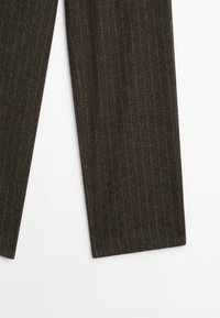 Jambes de pantalon en tissu marron foncé à fines rayures sur fond blanc, montrant une coupe droite et des lignes verticales fines.