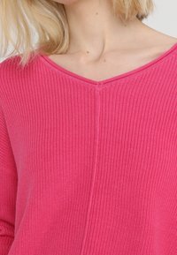 Sweter z dekoltem w kształcie litery V, z długim rękawem, w intensywnym różowym kolorze, wykonany z fakturowanej dzianiny. Posiada centralny szew oraz ściągacz przy dekolcie i na dole.