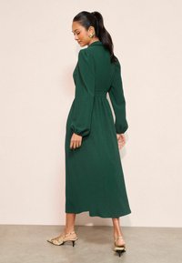 Gara zaļa kleita ar teksturētu audumu, pieguļošu vidukli un pūstajām piedurknēm. Valkāta kopā ar bēša krāsas apaviem ar čūskas ādas rakstu, uzsverot elegantu siluetu.