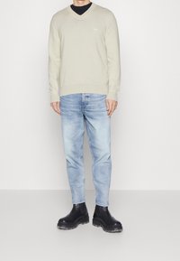 Beżowy sweter z dekoltem w serek na czarnej koszuli, w połączeniu z jasnoniebieskimi jeansami i czarnymi masywnymi butami. Sweter ma subtelne logo na piersi.