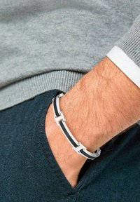 Bracciale in bianco e nero realizzato in metallo e gomma, con maglie e dettagli, indossato su un polso sopra un maglione grigio e un tessuto scuro.