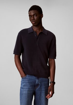 Mladi muškarac koji nosi crnu pletenu polo majicu kratkih rukava i plave traperice, stoji s jednom rukom u džepu nasuprot jednostavne sive pozadine.