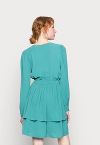 Vestido teal com mangas longas, cintura franzida e saia em camadas. Tecido suave com uma textura subtil, decote redondo e cintura elástica.