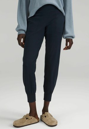 Persoon draagt marineblauwe joggingbroek, lichtblauwe geribde trui en beige suède slip-on klompen, staand op een effen achtergrond.