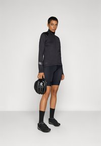 Black cycling jacket features high collar, full zip, and reflective accents. Paired with black shorts, socks, and helmet. Footwear is black cycling shoes.
