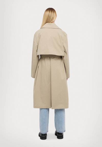 Beige trench coat, hátul rövidre szabva, hosszú ujjakkal és megkötött derékkal. Az anyag simának tűnik, enyhe fényességgel. Világoskék farmernadrággal párosítva.